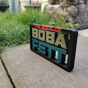 Peut inclure: Un panneau noir encadr&eacute; avec le texte "The Book of Boba Fett" en lettres en relief multicolores. Les lettres sont vertes, rouges et bleues.