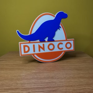 Może przedstawiać: Sylwetka niebieskiego dinozaura w białym owalu z pomarańczowym kółkiem i napisem "DINOCO" białymi literami.