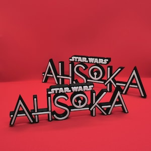 May include: Black and white "Star Wars Ahsoka" logo. The word "Ahsoka" is in a bold, stylized font with white outlines. The "Star Wars" text is above the name. The logos are on a red background.