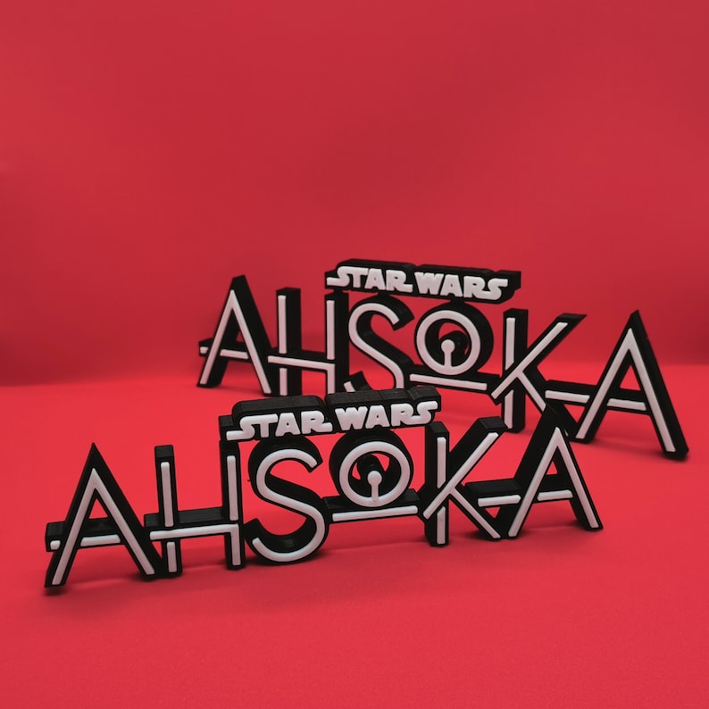 May include: Black and white "Star Wars Ahsoka" logo. The word "Ahsoka" is in a bold, stylized font with white outlines. The "Star Wars" text is above the name. The logos are on a red background.