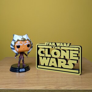 May include: A Star Wars: The Clone Wars logo with a black background and yellow lettering. A Funko Pop! figure of Ahsoka Tano stands in front of the logo.
