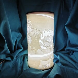 May include: A white ceramic lamp with a cylindrical shape features an embossed design of two Star Wars characters, Ahsoka Tano and Anakin Skywalker. The lamp is illuminated from within, casting a soft glow on the characters.