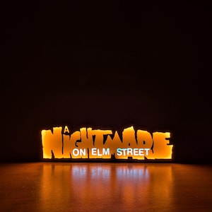 May include: Illuminated sign with the text "A Nightmare on Elm Street" in a stylized, orange font. The words "ON ELM STREET" are in a smaller, white font. The sign is lit from behind, creating a warm glow.