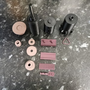 May include: A set of 3D printed parts in black and brown. The set includes a tall cylinder, two smaller cylinders, a small ring, a rectangular block, a small rectangle, a small circle, a small oval, and a small piece with a series of holes.