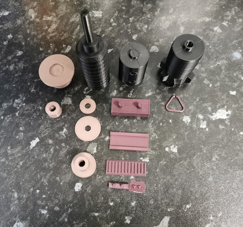 May include: A set of 3D printed parts in black and brown. The set includes a tall cylinder, two smaller cylinders, a small ring, a rectangular block, a small rectangle, a small circle, a small oval, and a small piece with a series of holes.