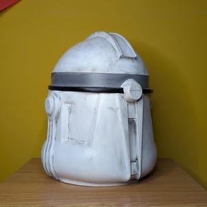 May include: A white and gray helmet with a rounded top and a dark gray band around the middle. The helmet has a weathered appearance and is likely a replica or prop.