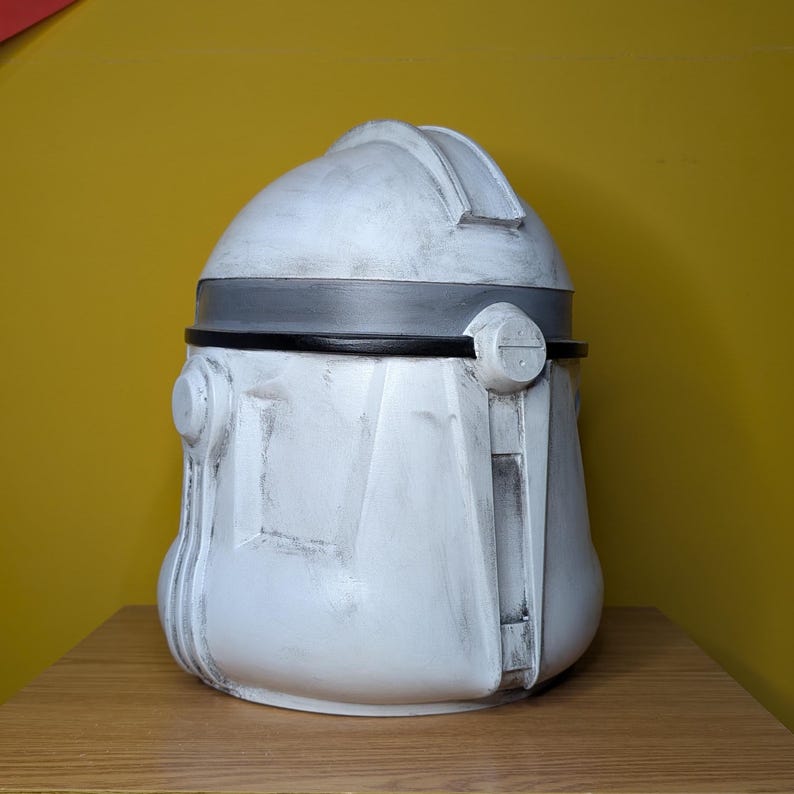 May include: A white and gray helmet with a rounded top and a dark gray band around the middle. The helmet has a weathered appearance and is likely a replica or prop.