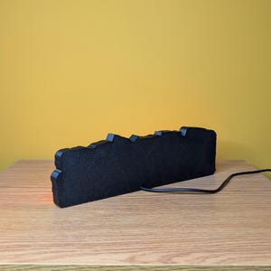 May include: A black, rectangular, decorative light with a jagged top edge. The light is on a wooden surface with a yellow background. A black power cord extends from the light.