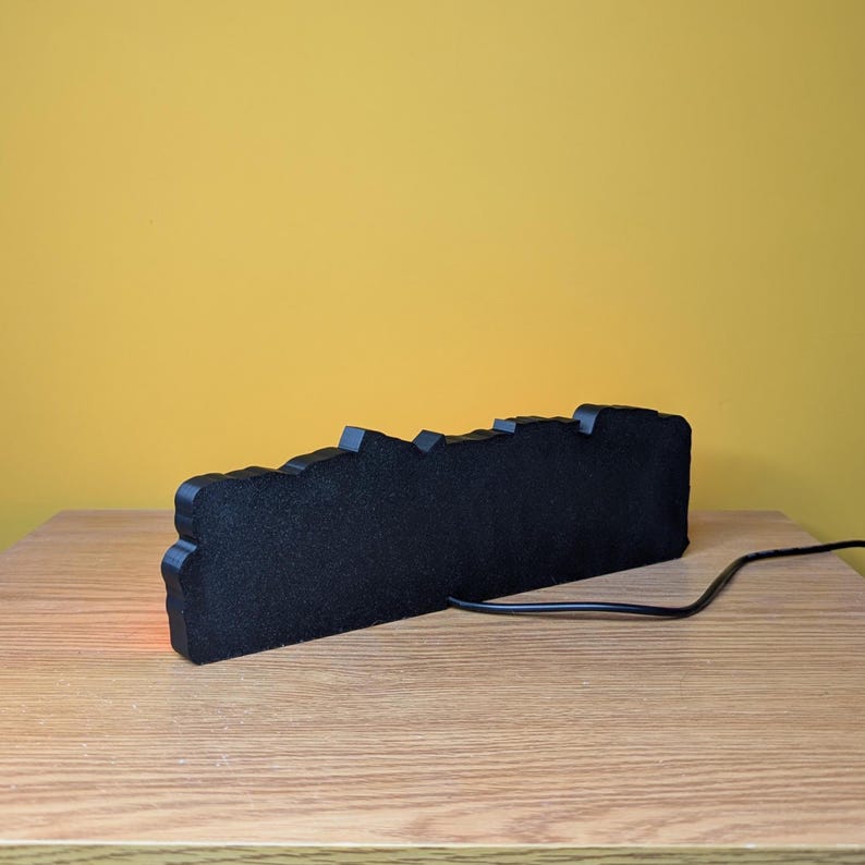 May include: A black, rectangular, decorative light with a jagged top edge. The light is on a wooden surface with a yellow background. A black power cord extends from the light.