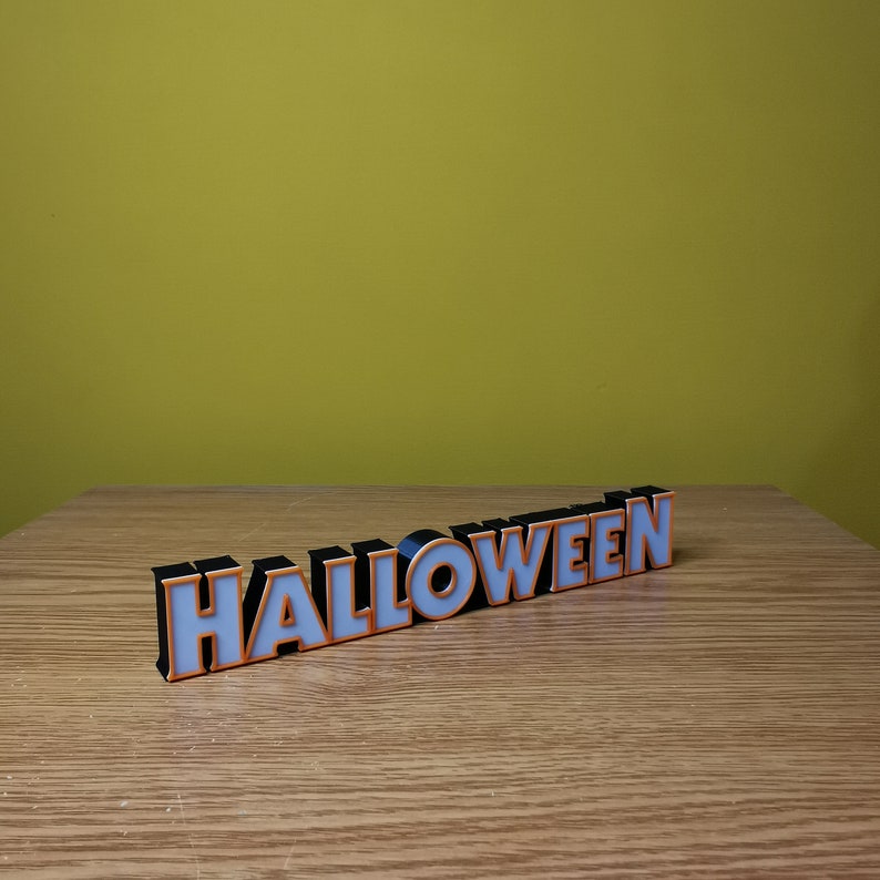 K&ouml;nnte beinhalten: Schwarze und wei&szlig;e Blockbuchstaben, die "HALLOWEEN" buchstabieren, mit orangefarbener Umrandung. Die Buchstaben liegen auf einer Holzoberfl&auml;che.