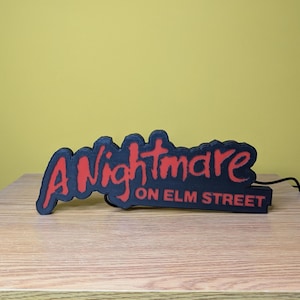 May include: Black and red sign with the text "A Nightmare on Elm Street". The sign is a 3D-printed design, with the words in a stylized font. The sign is placed on a wooden surface.