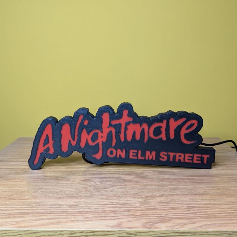 May include: Black and red sign with the text "A Nightmare on Elm Street". The sign is a 3D-printed design, with the words in a stylized font. The sign is placed on a wooden surface.