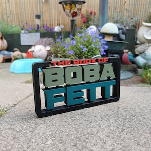 Peut inclure: Une plaque noire en 3D avec le texte "THE BOOK OF BOBA FETT" en rouge, vert et bleu. La plaque est pos&eacute;e sur une surface en b&eacute;ton gris.
