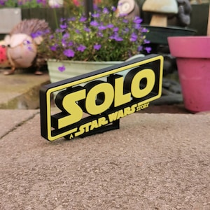 May include: A black and yellow 3D sign with the word "SOLO" in yellow letters and the text "A STAR WARS STORY" in smaller black letters.