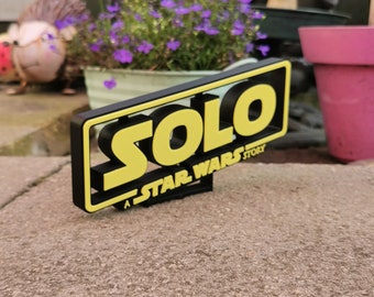 May include: A black and yellow 3D sign with the word "SOLO" in yellow letters and the text "A STAR WARS STORY" in smaller black letters.
