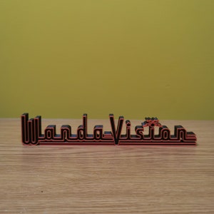 Peut inclure: Panneau imprim&eacute; en 3D noir et rouge avec le texte "WandaVision". Le texte est dans une police stylis&eacute;e. Le panneau est pos&eacute; sur une surface en bois, avec un fond jaune-vert.