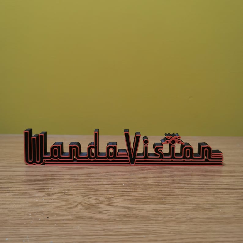Peut inclure: Panneau imprim&eacute; en 3D noir et rouge avec le texte "WandaVision". Le texte est dans une police stylis&eacute;e. Le panneau est pos&eacute; sur une surface en bois, avec un fond jaune-vert.