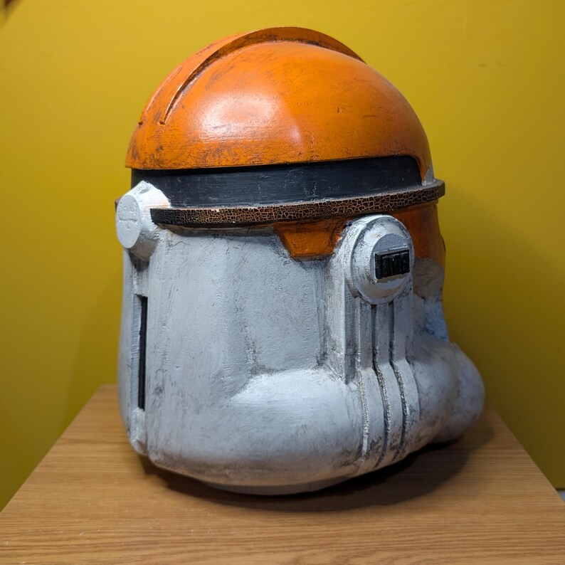 May include: A white and orange plastic helmet with a black visor. The helmet has a textured surface and is designed to look like a Star Wars clone trooper helmet.