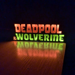 Pu&ograve; includere: Insegna luminosa con le parole "DEADPOOL" in arancione e "WOLVERINE" in verde. L'insegna si riflette su una superficie di legno. Lo sfondo &egrave; scuro.