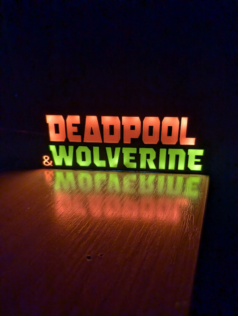Pu&ograve; includere: Insegna luminosa con le parole "DEADPOOL" in arancione e "WOLVERINE" in verde. L'insegna si riflette su una superficie di legno. Lo sfondo &egrave; scuro.