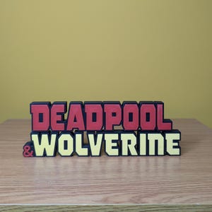 Könnte beinhalten: Ein 3D-gedrucktes Schild mit den Worten "DEADPOOL & WOLVERINE" in schwarzen, roten und gelben Buchstaben.
