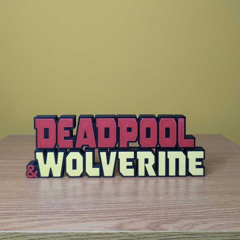 Może przedstawiać: Tr&oacute;jwymiarowy znak z napisem "DEADPOOL & WOLVERINE" w czarnych, czerwonych i ż&oacute;łtych literach.