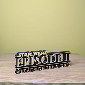 May include: A black and gold 3D printed sign that reads "Star Wars Episode II Attack of the Clones".