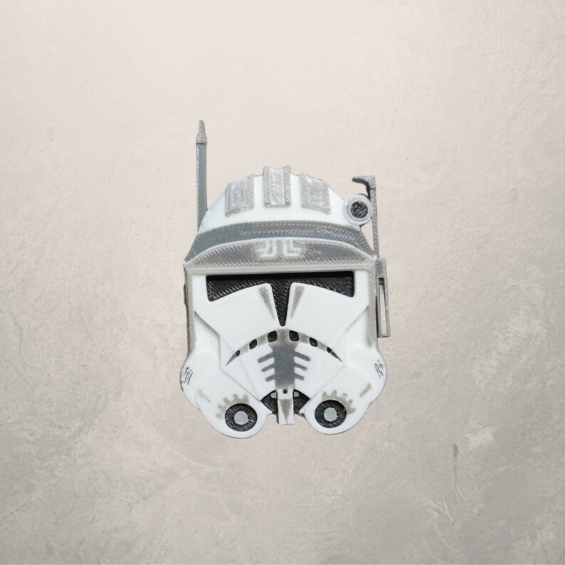 May include: A white and gray plastic clone trooper helmet with a black visor and a gray antenna. The helmet has a gray design on the front and a gray symbol on the top.
