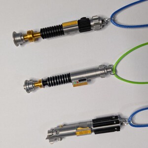 May include: Three miniature lightsaber pendants with silver, black, and gold details, each attached to a different colored cord: blue, green, and blue. The lightsabers have intricate designs and are displayed against a white background.
