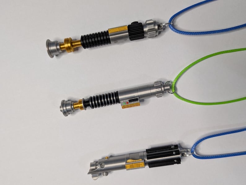 May include: Three miniature lightsaber pendants with silver, black, and gold details, each attached to a different colored cord: blue, green, and blue. The lightsabers have intricate designs and are displayed against a white background.