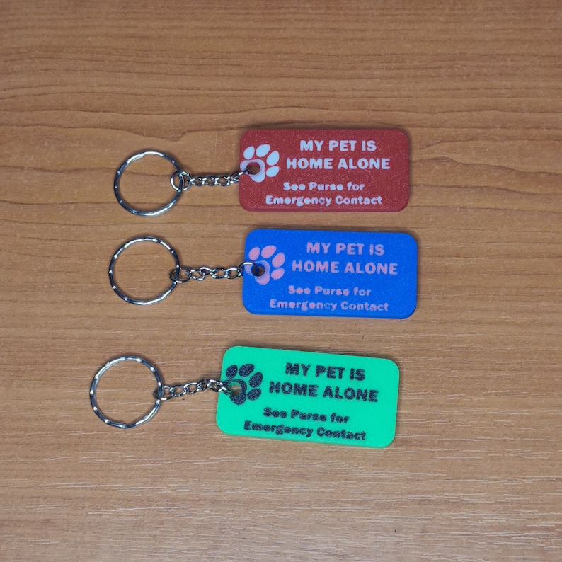 May include: Three keychains with rectangular tags that say "My Pet is Home Alone" in black text. The tags are red, blue, and green. Each tag has a paw print with a white outline.