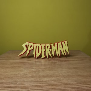 May include: A yellow and red 3D word sign that reads "SPIDER-MAN". The sign is on a wooden surface.