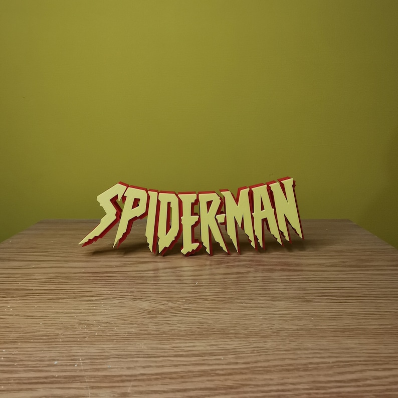May include: A yellow and red 3D word sign that reads "SPIDER-MAN". The sign is on a wooden surface.