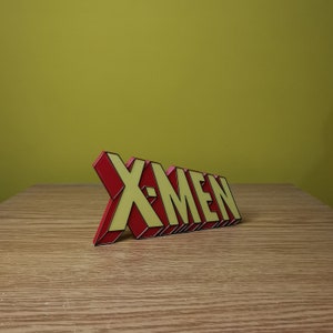 K&ouml;nnte beinhalten: Ein rotes und gelbes 3D-Plastikschild mit der Aufschrift "X-MEN" in einer fetten Comic-Schriftart. Das Schild steht auf einer Holzoberfl&auml;che.