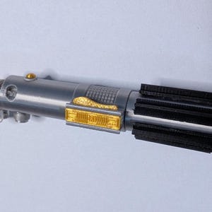May include: A silver and black lightsaber keychain with gold accents. The keychain features a detailed replica of a lightsaber, with a silver handle, black emitter, and gold detailing. A blue key ring is attached.