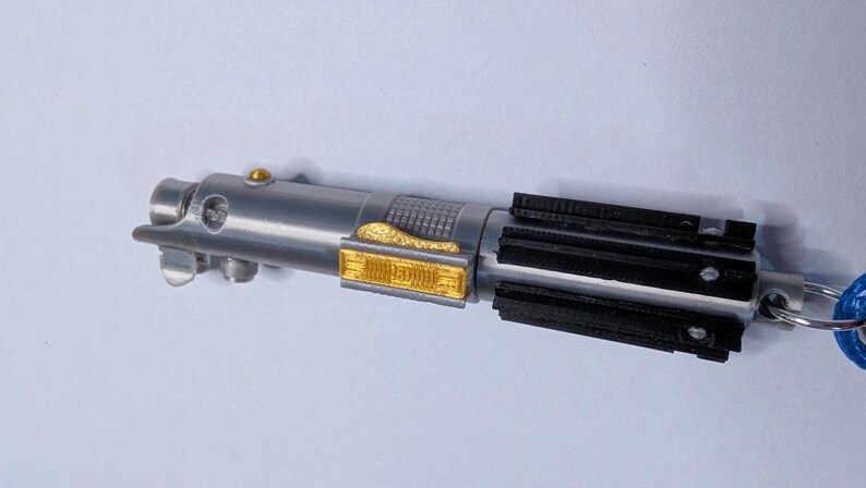 May include: A silver and black lightsaber keychain with gold accents. The keychain features a detailed replica of a lightsaber, with a silver handle, black emitter, and gold detailing. A blue key ring is attached.
