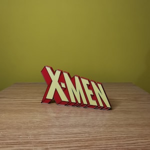 K&ouml;nnte beinhalten: Ein rotes und gelbes 3D-Plastikschild mit der Aufschrift "X-MEN" in schwarzer Umrandung. Das Schild steht auf einer Holzoberfl&auml;che.