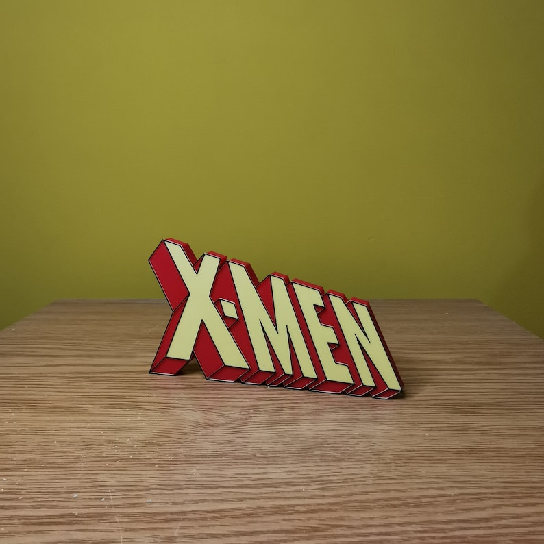 K&ouml;nnte beinhalten: Ein rotes und gelbes 3D-Plastikschild mit der Aufschrift "X-MEN" in schwarzer Umrandung. Das Schild steht auf einer Holzoberfl&auml;che.