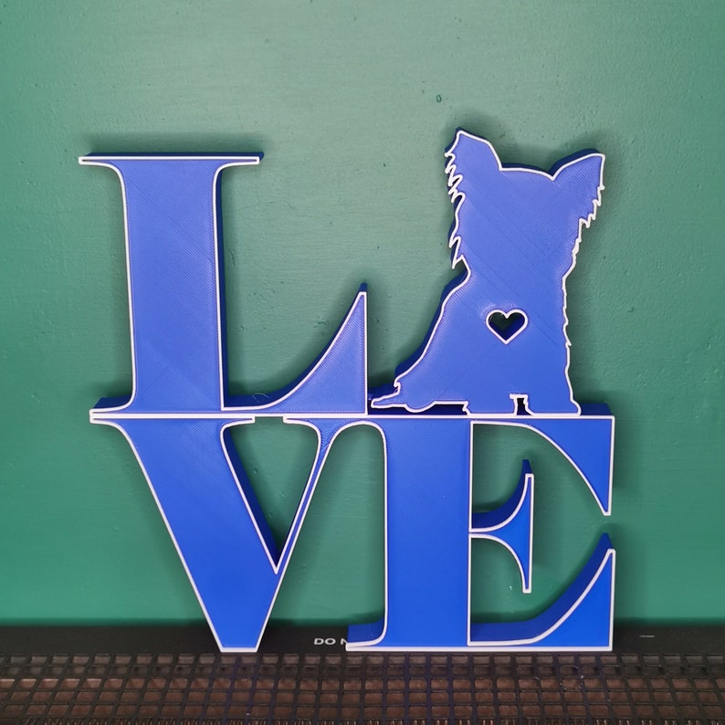 May include: Blue wooden cutout of the word "LOVE" with a white outline. A white silhouette of a dog is positioned on top of the "O" in the word.