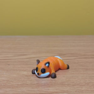 May include: A small orange and brown figurine of a fox lying on its side with its eyes closed. The fox has a white belly and a black nose.