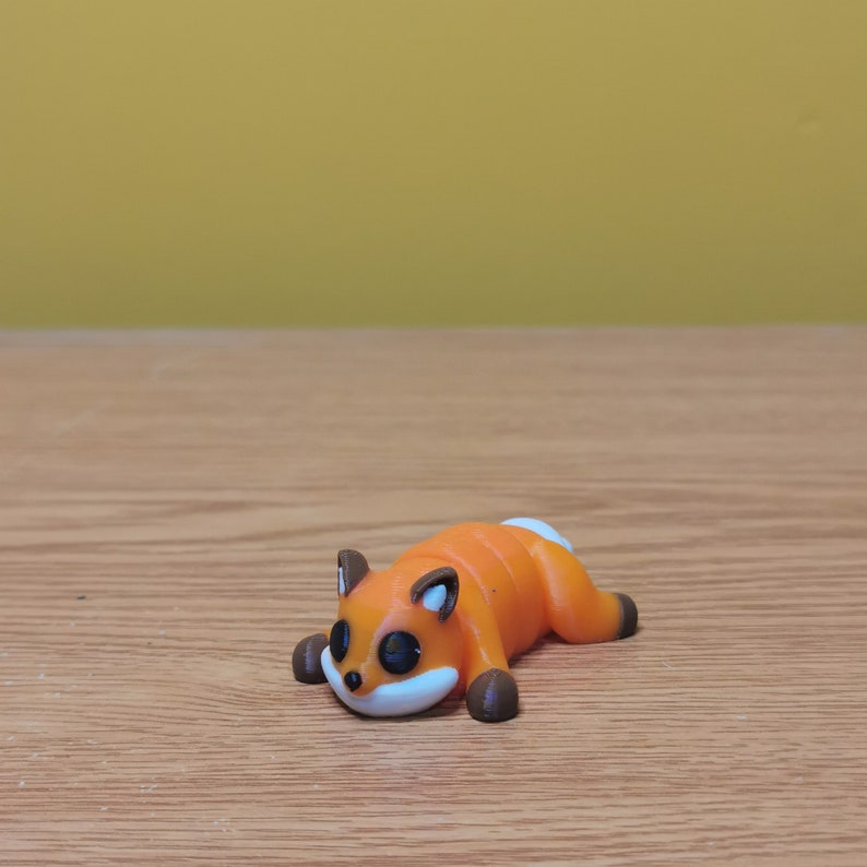 May include: A small orange and brown figurine of a fox lying on its side with its eyes closed. The fox has a white belly and a black nose.