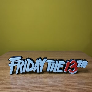 Puede incluir: Letras impresas en 3D en blanco y negro que deletrean "Viernes 13" con un "13" rojo que gotea sobre una superficie de madera.