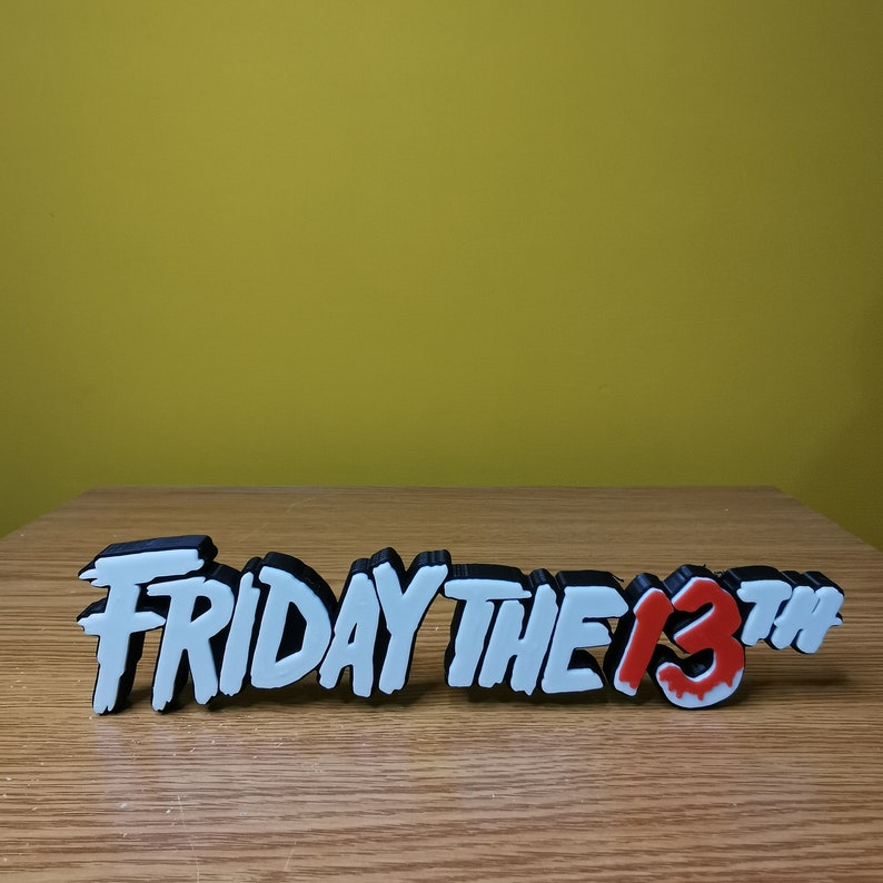 Puede incluir: Letras impresas en 3D en blanco y negro que deletrean "Viernes 13" con un "13" rojo que gotea sobre una superficie de madera.