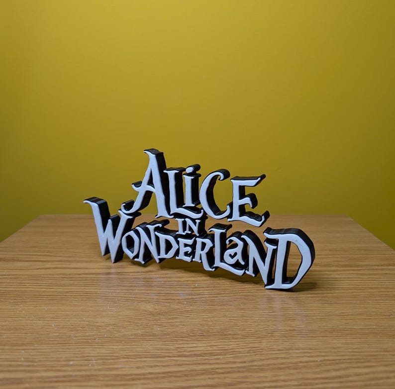 Pode incluir: Uma placa decorativa em preto e branco com o texto "Alice in Wonderland". A placa &eacute; feita de um material s&oacute;lido e &eacute; colocada sobre uma superf&iacute;cie de madeira. O fundo &eacute; amarelo.