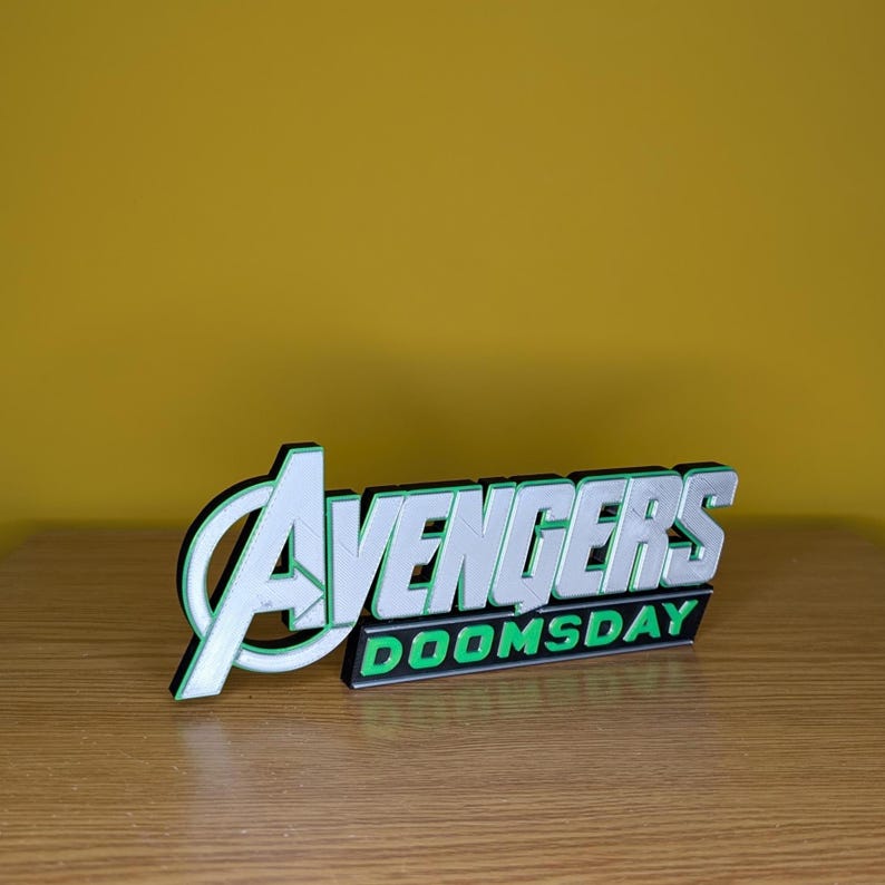以下が含まれることがあります： アベンジャーズのロゴと「DOOMSDAY」の文字が入った3Dプリントのサイン。白と緑の配色で、黒いアウトラインがあります。木製の表面に、黄色の背景に表示されています。