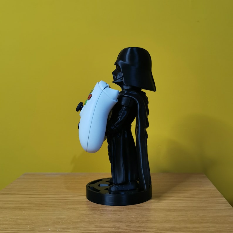 May include: A black Darth Vader figurine holding a white Xbox controller. The figurine is standing on a black base.