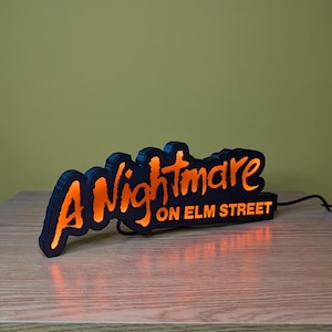May include: Black and orange illuminated sign that reads "A Nightmare on Elm Street." The sign has a black border and the text glows orange. The sign is plugged into an electrical outlet.