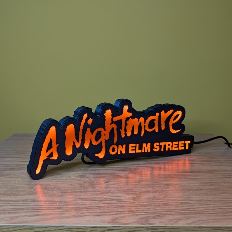 May include: Black and orange illuminated sign that reads "A Nightmare on Elm Street." The sign has a black border and the text glows orange. The sign is plugged into an electrical outlet.