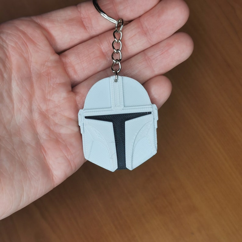 May include: White plastic keychain featuring a Mandalorian helmet design. The helmet is detailed with black accents and a visor.