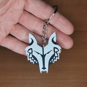 May include: A white wolf head keychain with black detailing. The keychain is attached to a silver chain.
